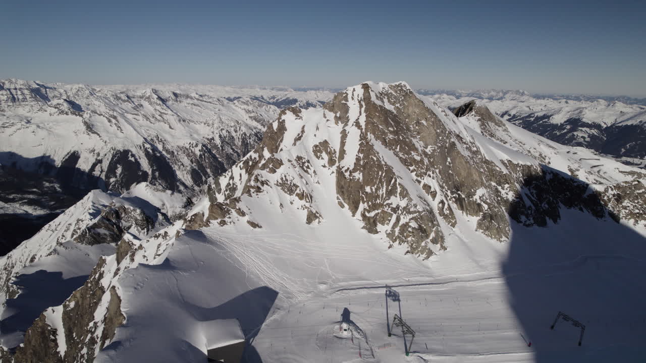 Aerial panning view of Sunny, Snowy Mountain Peak with Ski Piste and Mountain Range