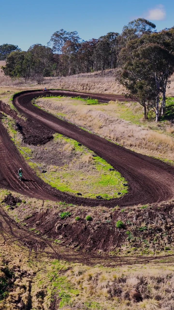 Un pilota di motocross sfreccia intorno a una curva di una pista sterrata nella rurale Tregony, Queensland, in pieno giorno, ripreso da una prospettiva aerea ampia e alta con drone.