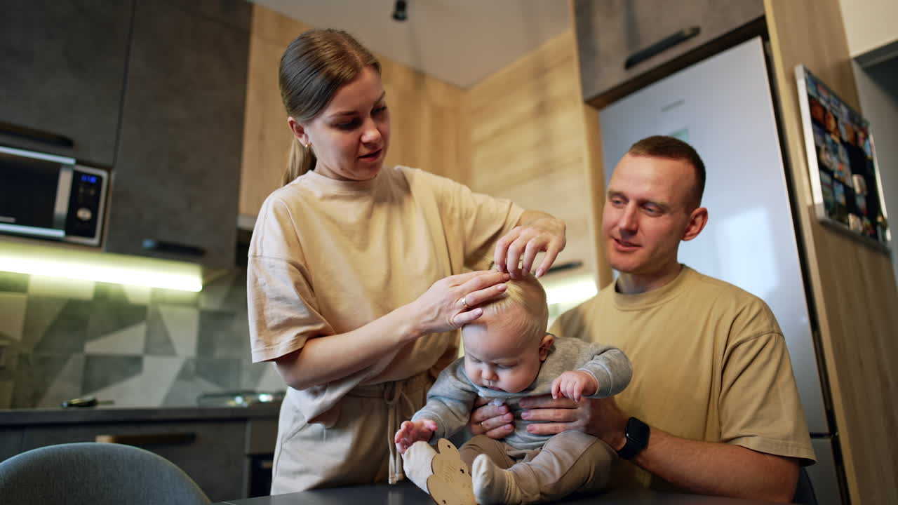 Father holding his cute baby boy sitting on the table. Mother ties a tiny pony tail on the blond hair of her son. Low angle view.