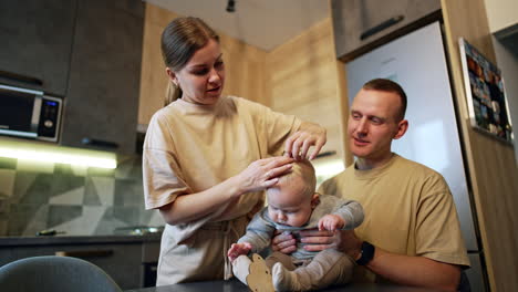 father holding his cute baby boy sitting on the table. mother ties a tiny pony tail on the blond hair of her son. low angle view.