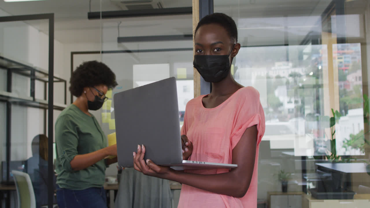 Happy african american businesswoman wearing face mask using laptop, looking to camera in office