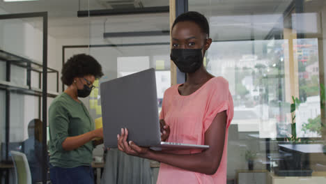 Feliz-Mujer-De-Negocios-Afroamericana-Usando-Mascarilla-Usando-Una-Computadora-Portátil,-Mirando-A-La-Cámara-En-La-Oficina