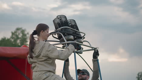 colleague working preflight setup as woman secures metal burner to balloon frame poles under overcast sky in grassy roadside field while partner adjusts support rods and gauges equipment