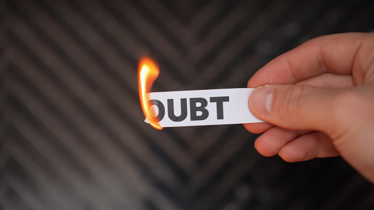 Close-up hand holds a paper strip reading “DOUBT” burning at the edge, symbolizing overcoming fear, self-belief, and mindset change with copy space on dark background