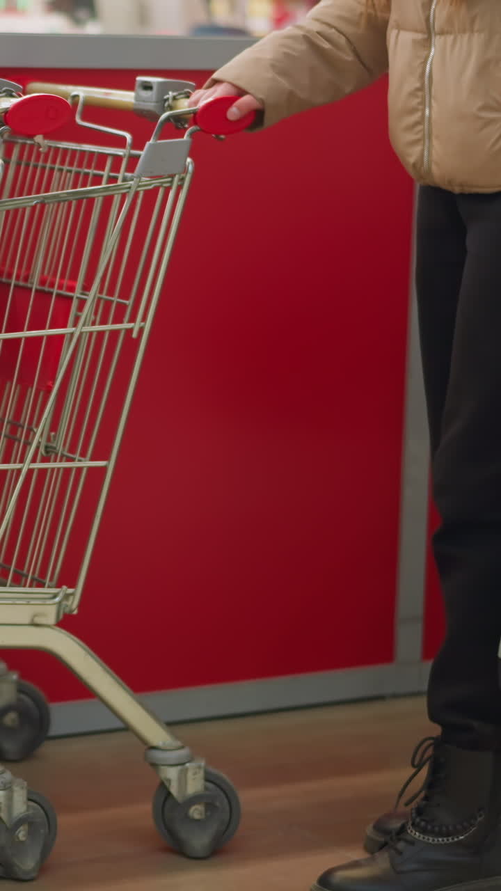 una mujer, con su cara fuera de la vista, se ve con una chaqueta de melocotón y pantalones negros mientras lleva una bolsa de gimnasia con correas rojas brillantes. alcanzando un carrito de compras en una tienda