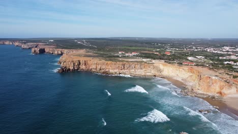 Droneshot-De-La-Hermosa-Playa-De-Tonel-Con-Grandes-Olas-Rodando-En-La-Bahía-Y-Sagres-En-Las-Colinas,-Sur-De-Portugal-Con-Clima-Soleado