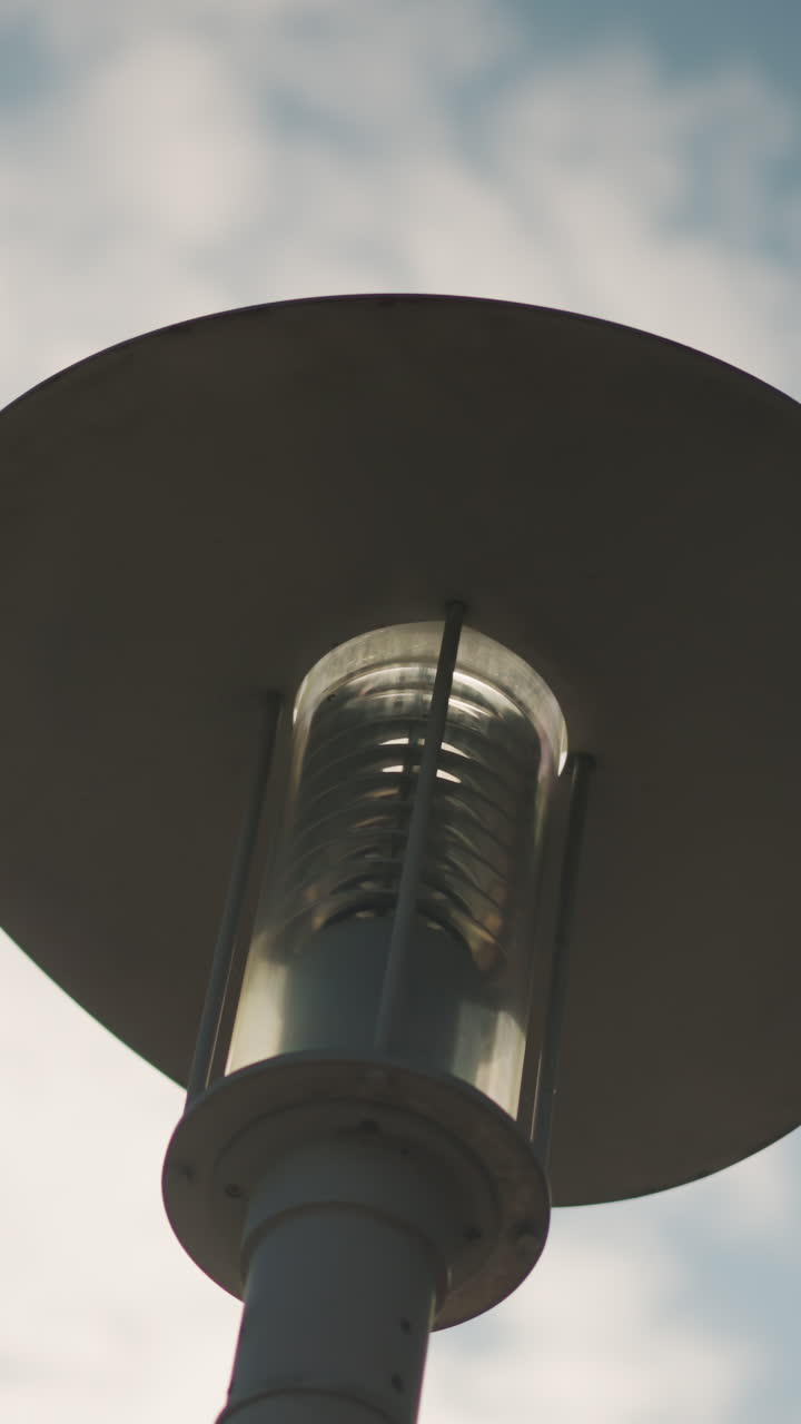 유리 실린더와 넓은 금속 갓이 있는 흐린 하늘 아래 가로등 실루엣, 은은한 햇빛과 부드러운 구름 배경이 몽환적인 한낮의 분위기를 연출하며 조명 디자인과 공공 장소를 위해 구도를 잡았어요.