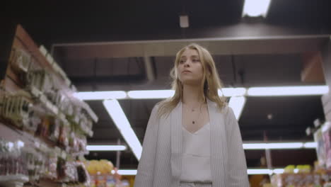 pretty lady in casual clothes is walking in grocery store steering shopping trolley with food inside it and looking around at shelves with products. women and shops concept.