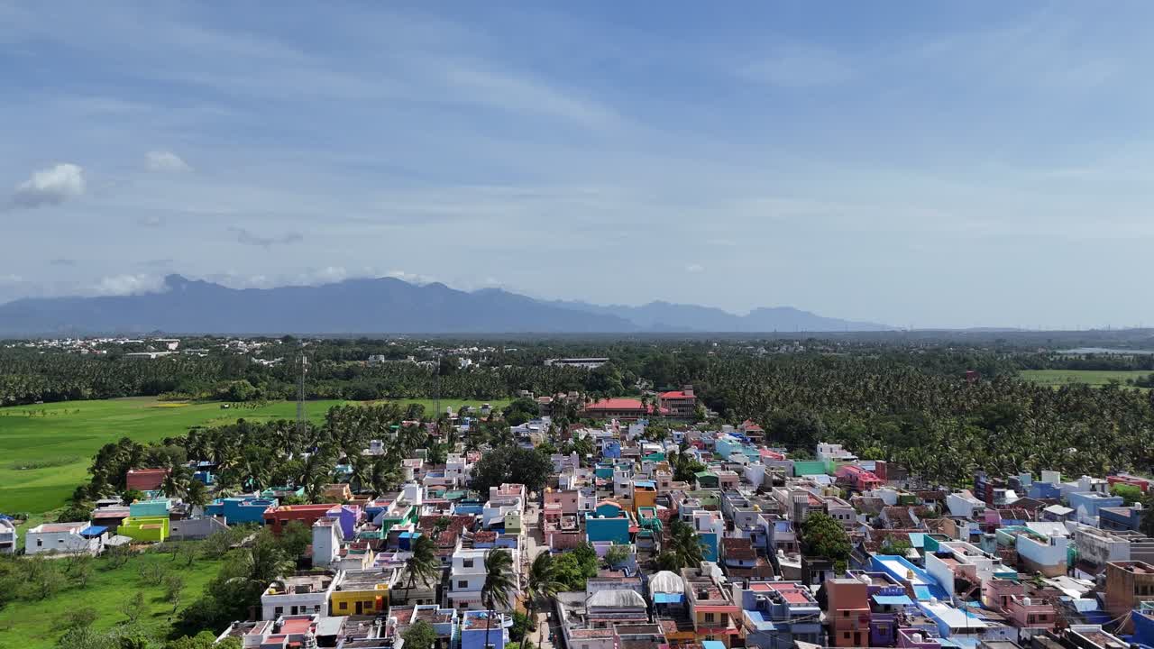 An aerial shot shows a vibrant village nestled at the foothills of the Western Ghats' majestic mountains. Colorful houses contrast with lush green forests and paddy fields under a bright blue sky