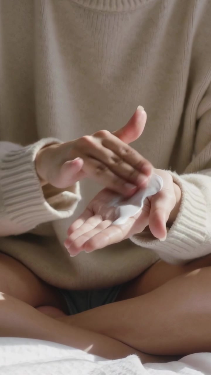 Vertical video: Noticing cream in left palm, woman in beige knit rubbing hands on bed for moisture