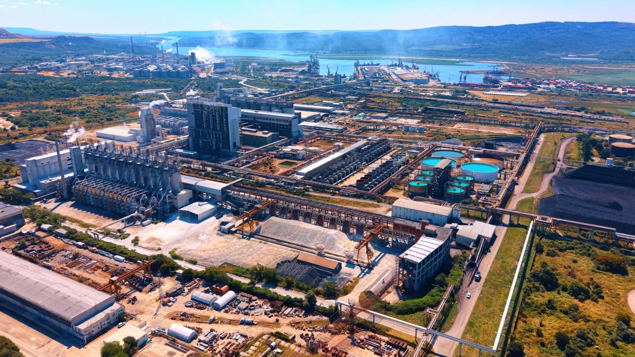 Panoramic view of large industrial complex near a waterway. Smoke rises from factory structures surrounded by storage yards, coal heaps, and distant hills.
