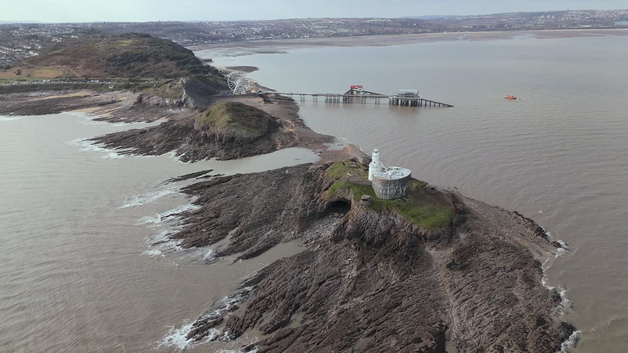 Moving drone view of Mumbles Lighthouse located on rocky terrain with The Big wheel at background during daytime in United Kingdom.