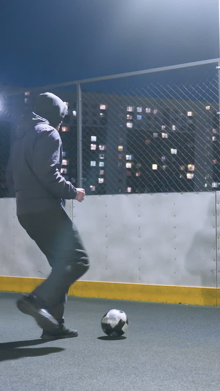 Man in hoodie kicking soccer ball against wall on illuminated urban sports field at night, with reflection visible on wall and city lights glowing in background