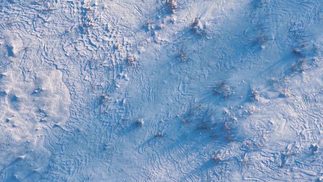 Drone video showcases intricate patterns on a snow-blanketed terrain, capturing untouched textures and subtle shadows in soft golden hour sunlight. No people or animals present.