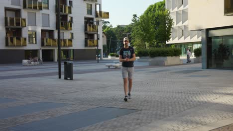 Male-Influencer-Vlogging-Using-Selfie-Stick-Walking-Around-Urban-Courtyard-Cityscape