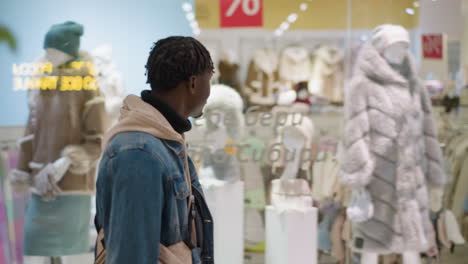 young black man in denim jacket and hoodie exploring winter coat mannequins in bright modern clothing store display thoughtful side profile shot capturing retail browsing urban shopper scene