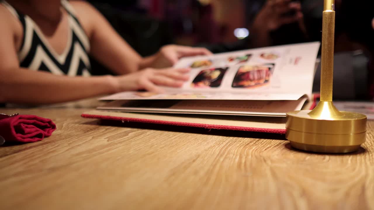 A woman in a patterned dress opens and flips through a restaurant menu at a warmly lit wooden table with a candle and rose, shot in Bangkok