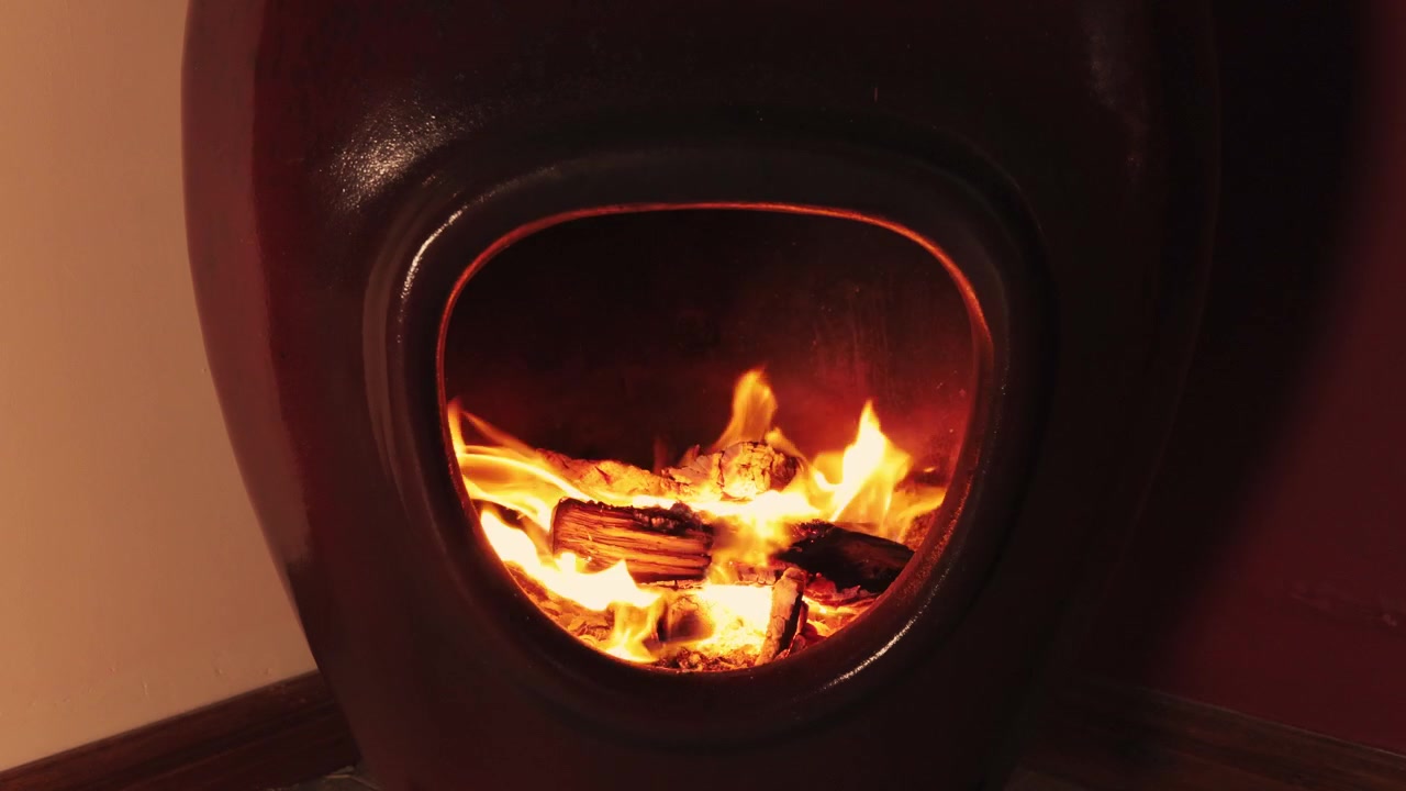 Bright orange flames consume firewood inside the smooth opening of a dark maroon ceramic freestanding fireplace. Fixed tripod shot. Indoor domestic setting with warm ambient light.