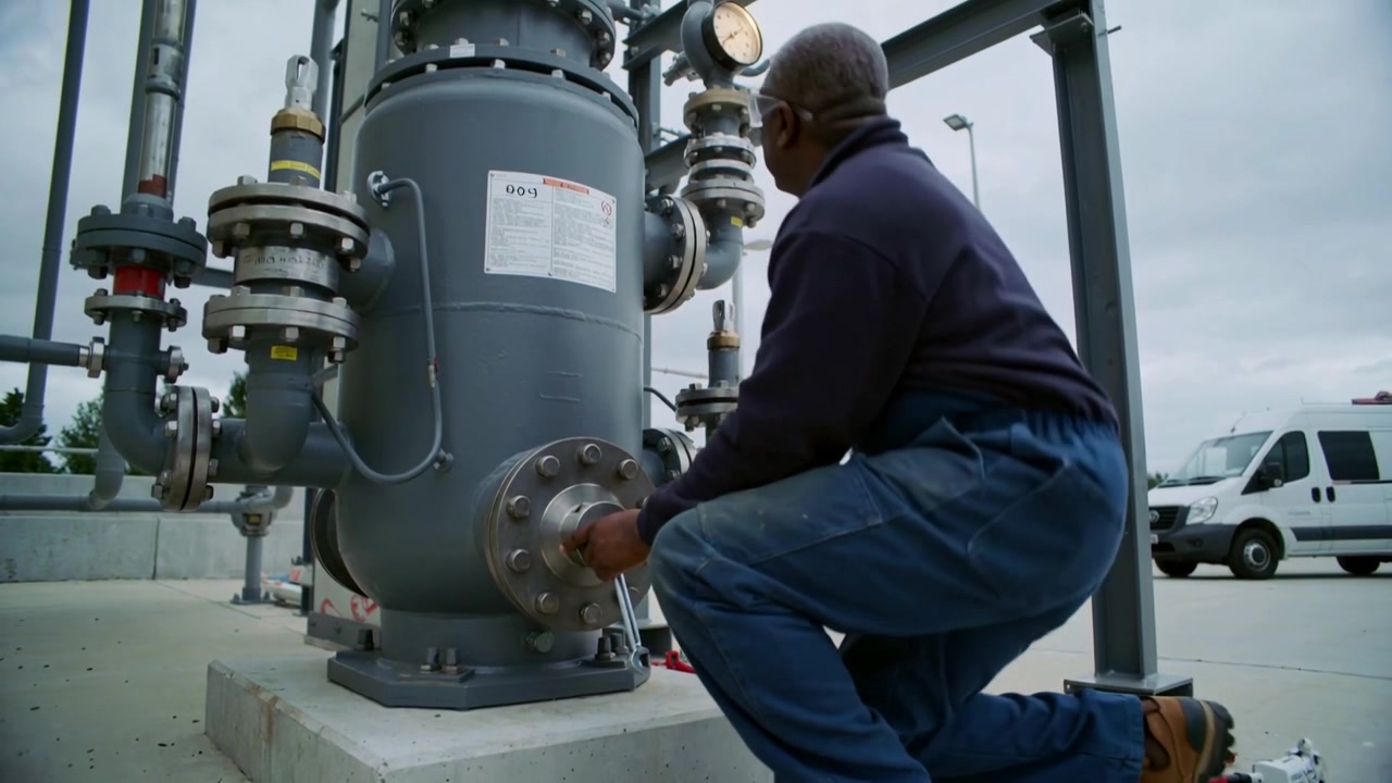 On call kneeling tech torquing flange with wrench at utility pad, wearing glasses, checking gauge