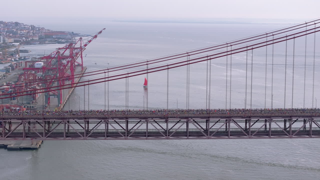 Side-on aerial drone shot of half marathon and 10K long-distance running event in Lisbon, Portugal, Europe. Runners crossing the iconic famous red 25th April bridge