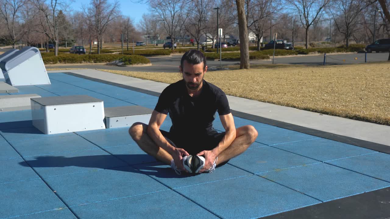 Hispanic man with beard does butterfly stretch at park outdoor fitness course