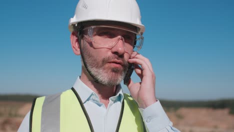 portrait sand quarry worker engineer, male caucasian foreman in safety wear and hardhat talks instructing builders with cellphone smartphone. builder, mining project specialist on sunny day