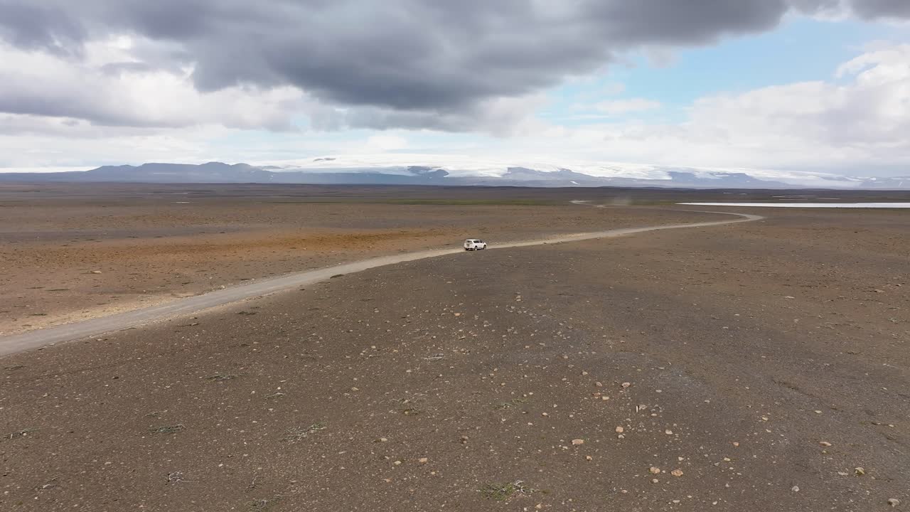 Aerial following shot of car on road in Icelandic landscape during cloudy day. Glacier mountains in background. Stony and rocky island of Iceland
