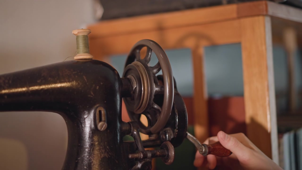 Primo piano di una macchina da cucire vintage, mano che infila l'ago e regola il volantino, sarta che restaura una macchina stile Singer in un accogliente laboratorio, la calda luce di una lampada evidenzia la patina metallica, rocchetto e bobina, con cura.