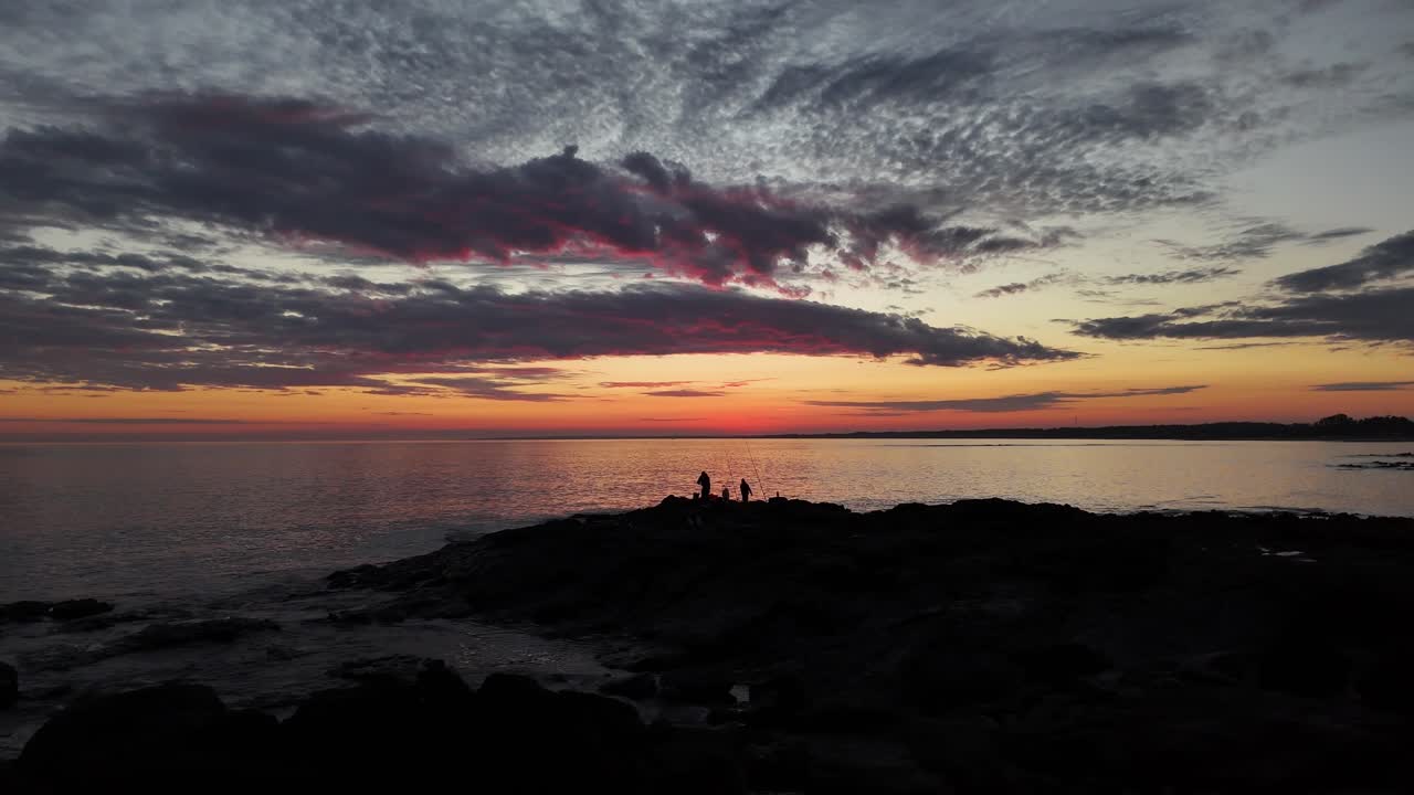 At Playa Verde, Uruguay, the drone orbits two fishermen silhouetted on rocks against vibrant post-sunset sky. Dramatic clouds and sea glow in magical twilight light