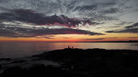 at playa verde, uruguay, the drone orbits two fishermen silhouetted on rocks against vibrant post-sunset sky. dramatic clouds and sea glow in magical twilight light