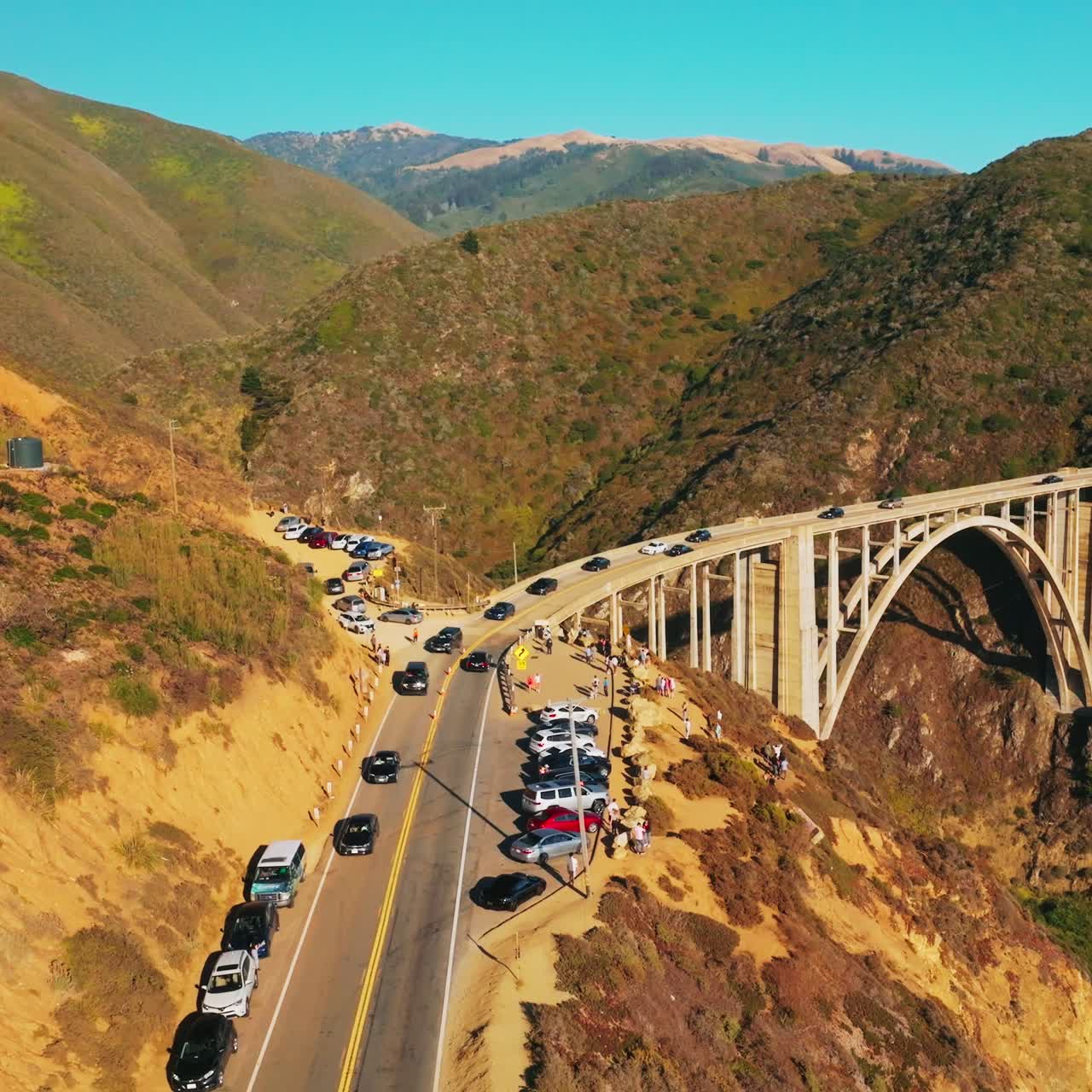 Cars moving slowly to and from the bridge in the mountains. California rocks at the coast of Pacific on sunny daytime. Aerial view