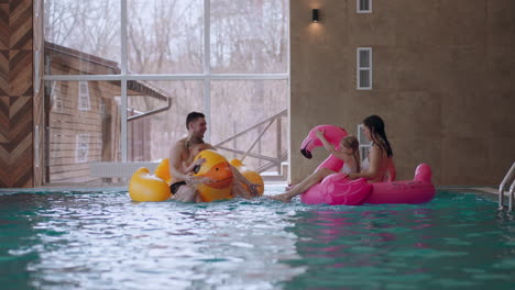 children and parents are resting in modern swimming pool indoors father and son are splashing mother and little girl