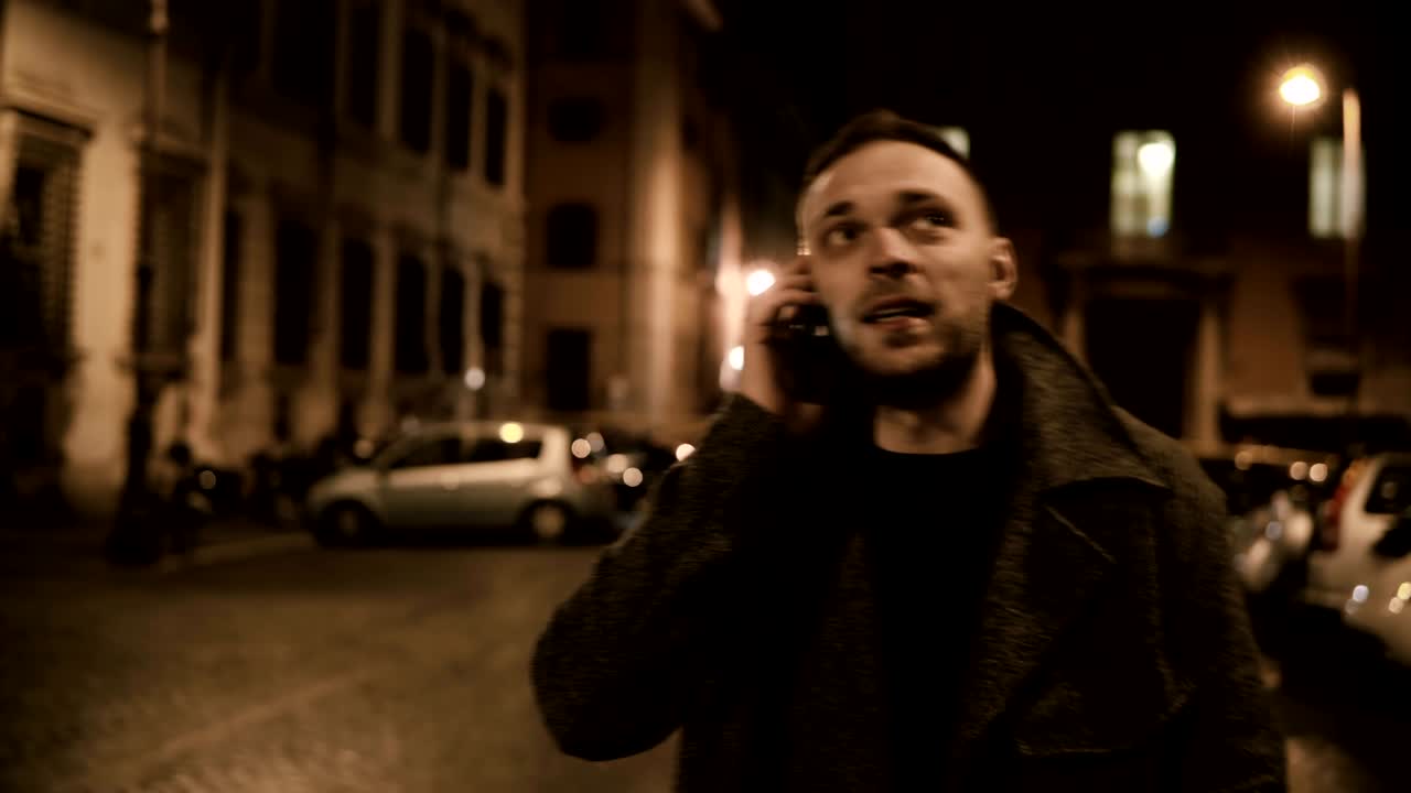 задний вид молодого красивого мужчины, идущего по улице вечером и использующего смартфон, разговаривающего с другом