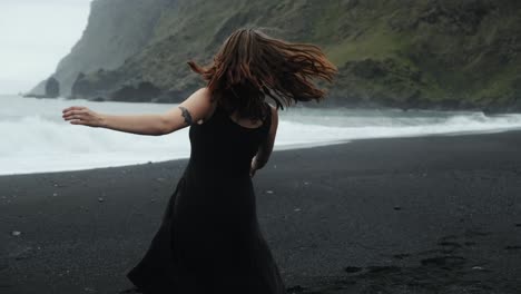 Junge-Rothaarige-Frau-In-Schwarzem-Kleid-Tanzt-Am-Schwarzen-Sandstrand-Islands,-Sich-Drehende-Zeitlupe,-Dramatische-Wellen-Meereslandschaft