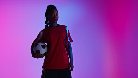 Studio-Portrait-Of-Female-Football-Or-Soccer-Player-Wearing-Team-Shirt-Holding-Ball-Under-Her-Arm-Shot-Low-Key-Against-Colourful-Mixed-Lighting