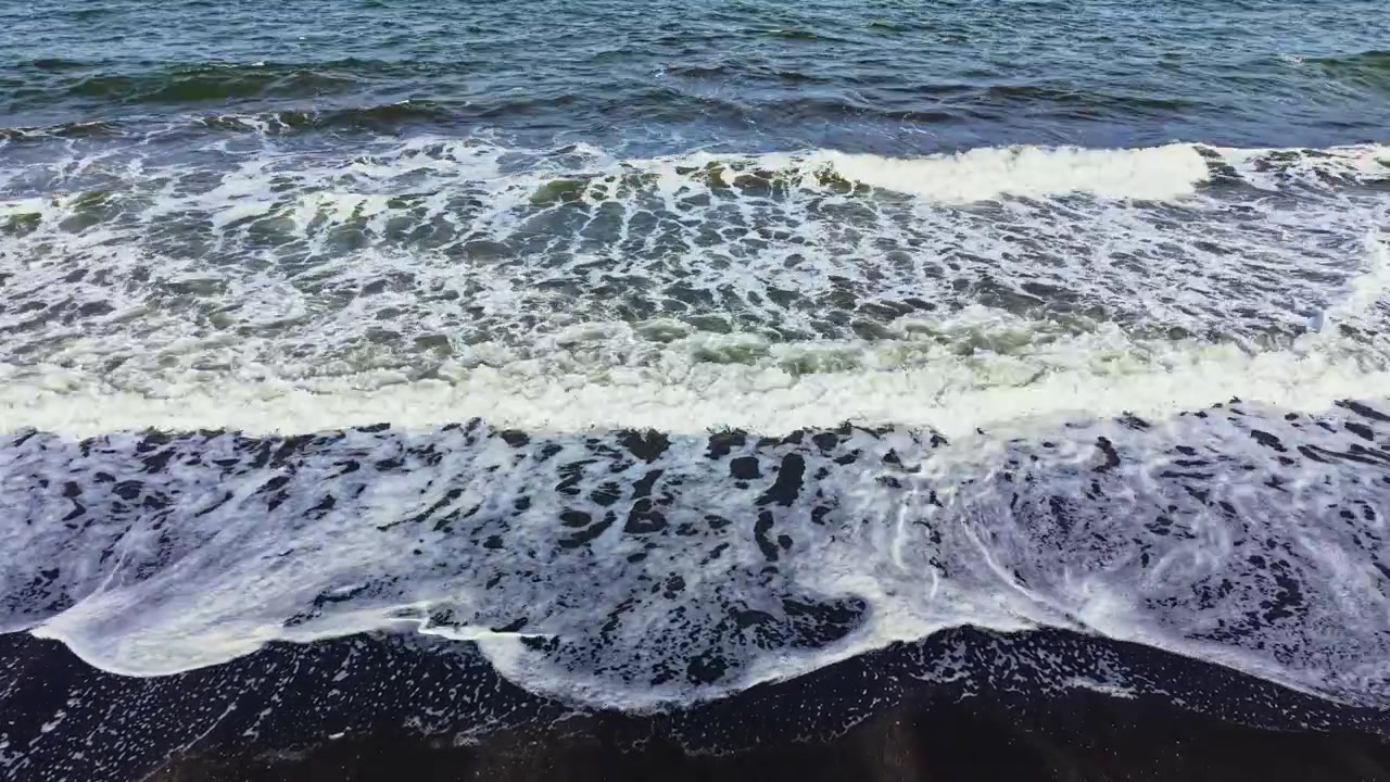 Le onde si infrangono su una spiaggia sabbiosa a mezzogiorno, creando schiuma e spruzzi lungo la riva mentre il sole splende luminoso in alto.