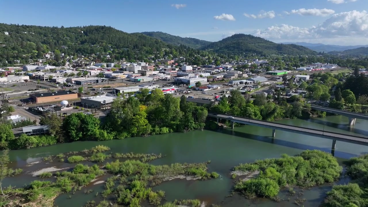 雲のある晴れた空の下、鬱蒼とした森の丘に囲まれた、高速道路の橋、商業ビル、並木道が特徴の緑豊かな川沿いのローズバーグのダウンタウン。アメリカ、オレゴン州。空撮ワイドショット。