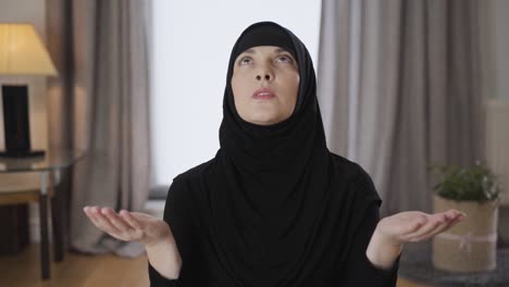 camera approaching slowly to attractive muslim woman in hijab praying at home. portrait of young lady in traditional hijab sitting with raised hands and talking. religion, traditional culture.