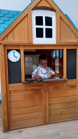 lovely toddler boy stands at the window of a fairy wooden playhouse. baby plays happily outdoors. vertical video.