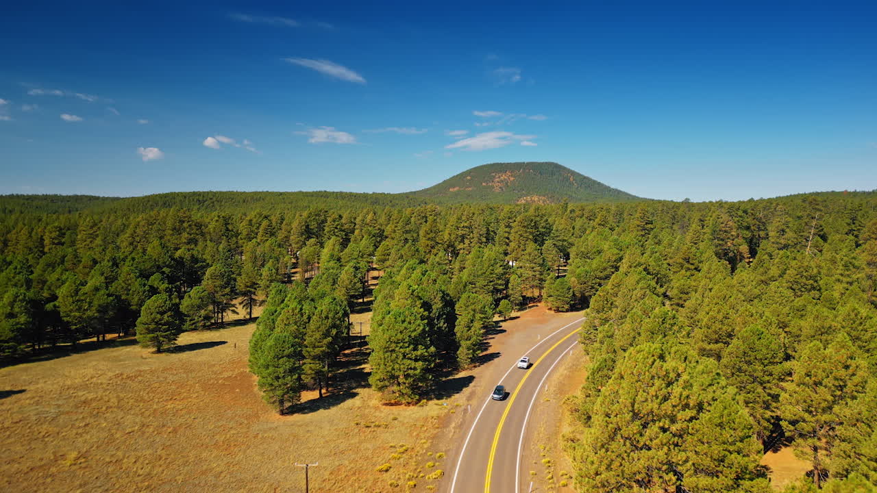 Two cars ride by highway in the woods. Verdant mountain against blue sky at backdrop. Aerial view