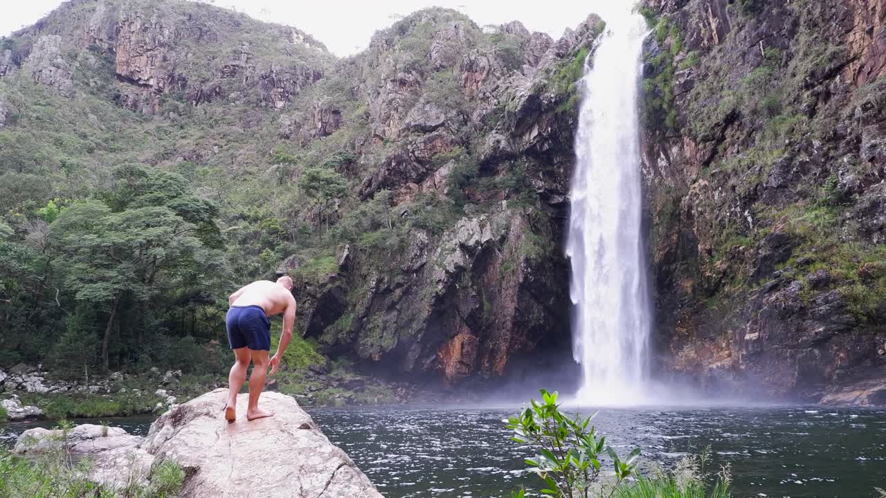 Caucasian male traveller in swim suit approaches vertical Waterfall