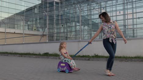 mother and daughter walking from airport after vacation. woman rides baby child on suitcase luggage