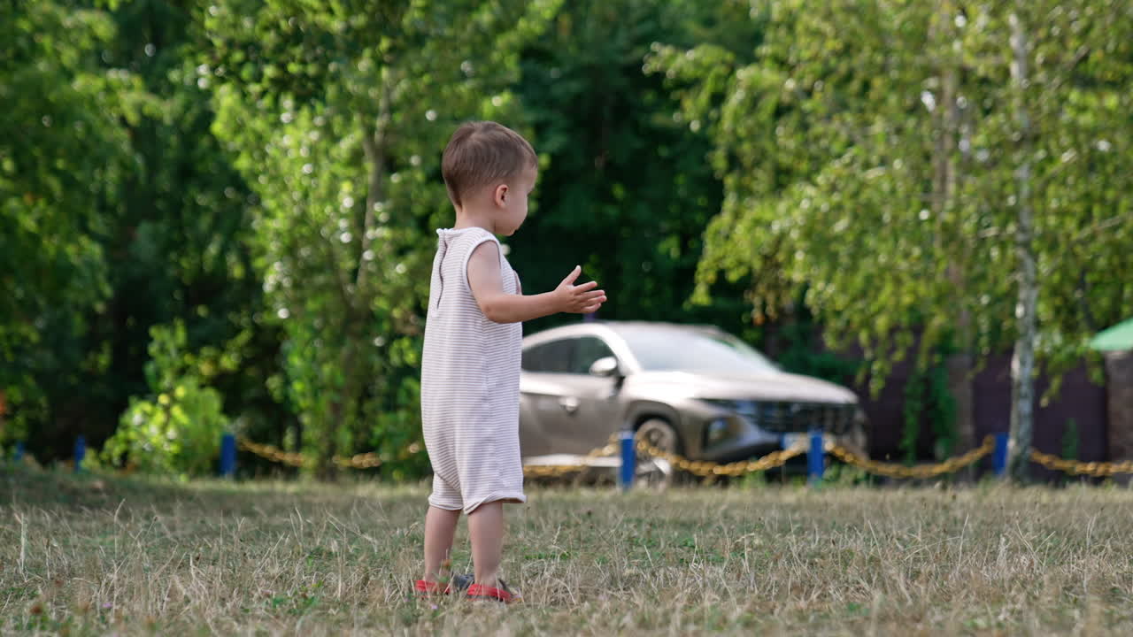 Caucasian baby in a bodysuit stands on the meadow. Toddler looks around and waves hands.