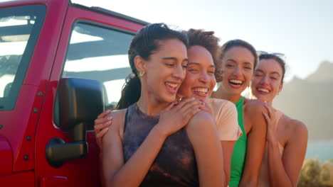 Portrait-Of-Laughing-Female-Friends-Standing-And-Hugging-By-Open-Top-Car-On-Road-Trip