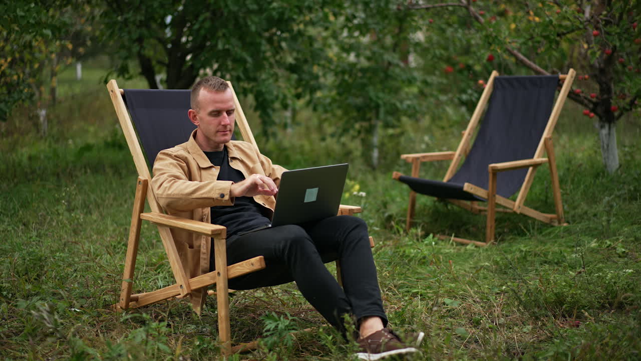 Caucasian male freelancer starting work on his computer. Man sits in a garden chair opening his laptop to start remote work. Nature backdrop.