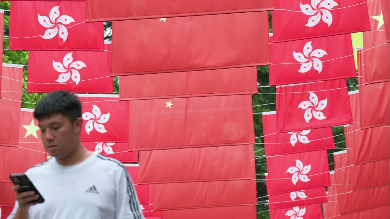 Low-angle view of flags of the People's Republic of China and Hong Kong SAR hang above a busy street as pedestrians walk below in Hong Kong, China.