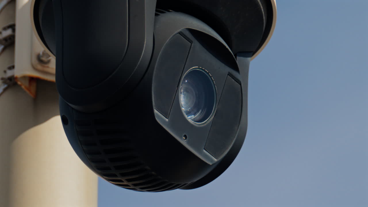 Close up of a round, black surveillance camera mounted on a white pole with the sky on the background