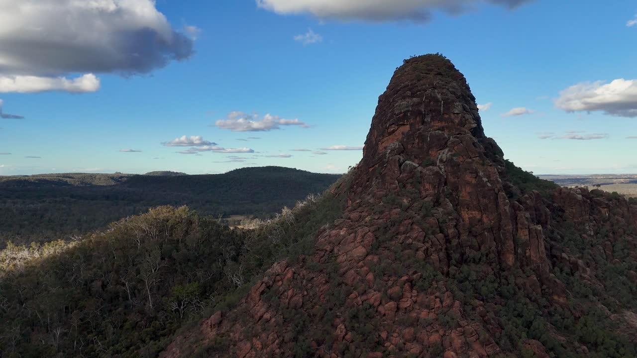 Drone camera smoothly circles Timor Rock, revealing rugged red sandstone, forested slopes, and expansive blue sky under soft natural daylight