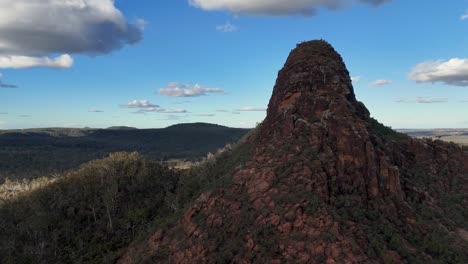 drone camera smoothly circles timor rock, revealing rugged red sandstone, forested slopes, and expansive blue sky under soft natural daylight