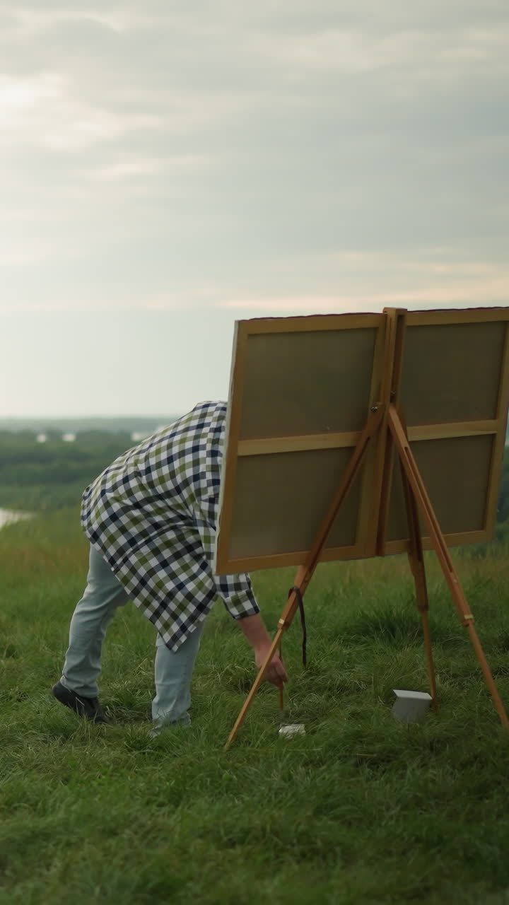 un artista con un sombrero negro, camisa a cuadros y vaqueros está pintando en un gran lienzo en un campo de hierba tranquila junto a un lago sereno